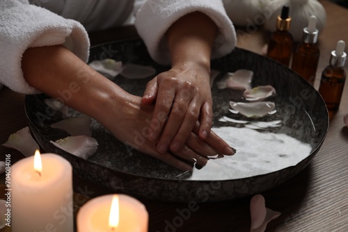 Woman soaking her hands in bowl of water and flower petals at wooden table, closeup. Spa treatment photo