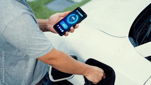 Hand insert EV charger to electric car to recharge battery while check battery status display on smartphone. Future sustainable and clean energy for EV car. Peruse © Summit Art Creations