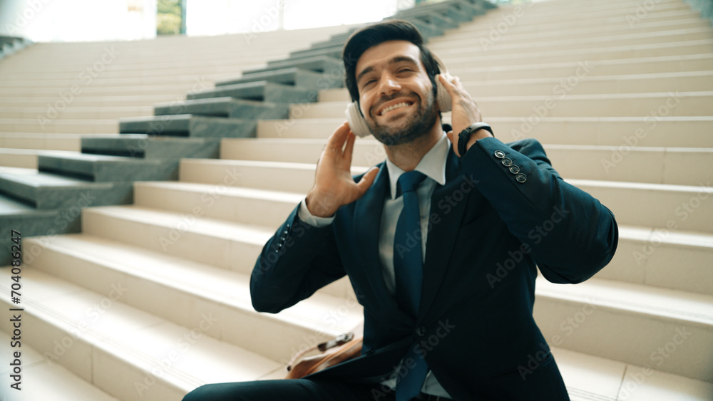 Project manager listening and enjoy music from headphone while sitting at stairs. Professional businessman wearing suit outfit while dancing and moving to music. Happy man listen funny song. Exultant. - obrazy, fototapety, plakaty 