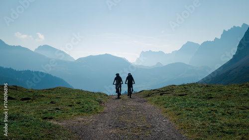 Silhouettes of two men mountainbiking in the Karwendel mountains in front of blue mountain layers during sunny blue sky day in summer, Tyrol Austria.