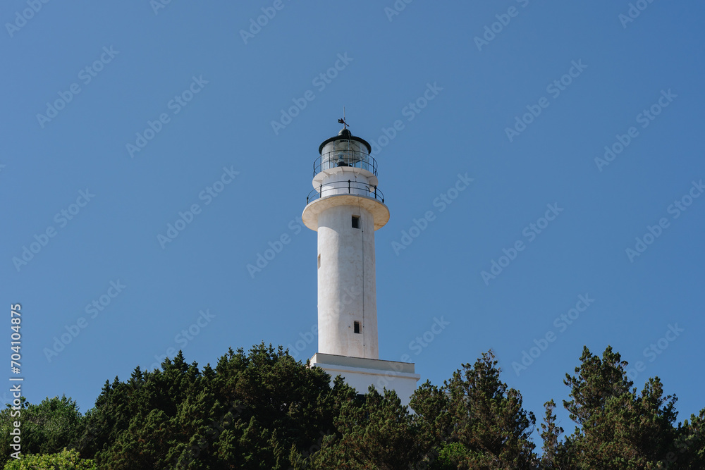 A white lighthouse stands tall above the trees against a clear blue sky, a beacon of navigation and hope