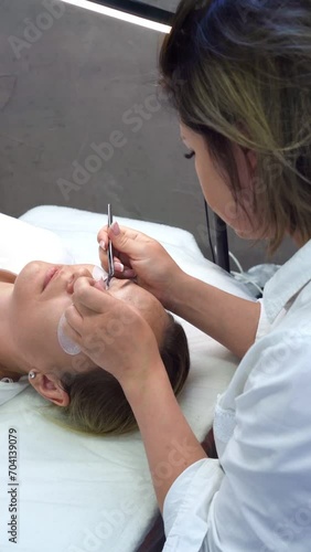 Video of young master sitting over client with patches on lower lids and checking durability of freshly extended eyelashes using tweezers. Woman touching black lashes with tool. Vertical.
