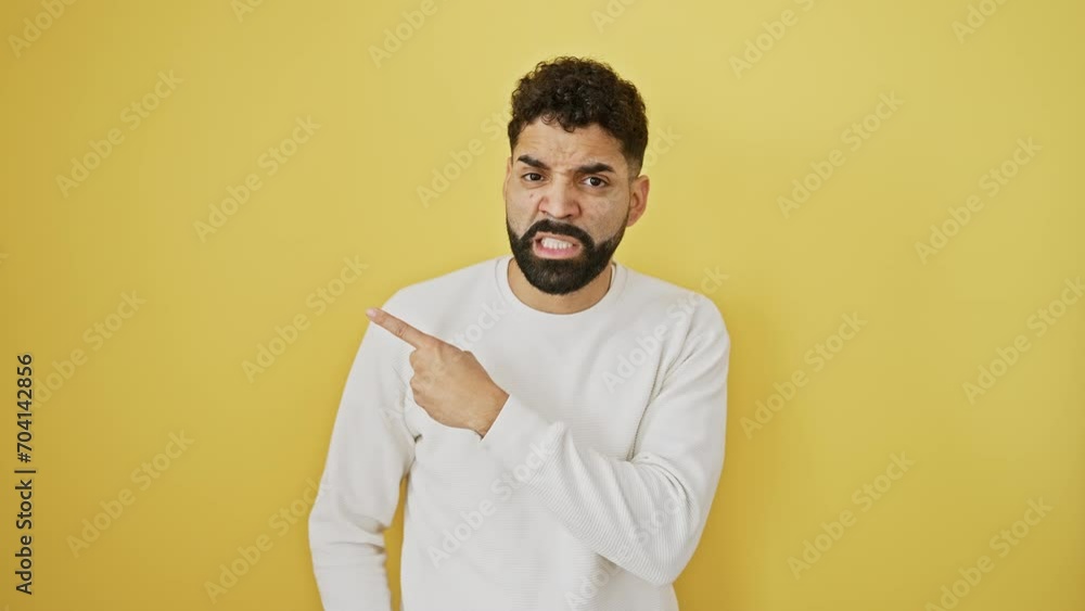 Worried young man, with a nervous expression, stands pointing aside. shock and surprise against a yellow background. his forefinger reveals his concern. Worried young man, with a nervous expression, stands pointing aside. shock and surprise against a yellow background. his forefinger reveals his concern.
