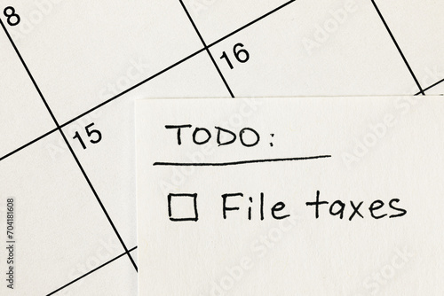 Hand written to-do list with 'File taxes' reminder written on a sticky note over a calendar, with income tax return filing deadline of April 15