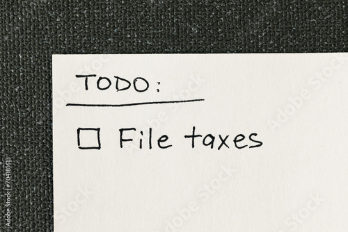 Handwritten to-do list with 'File taxes' reminder and a checkbox written on a white note against a gray textured backdrop, flat lay top-down view