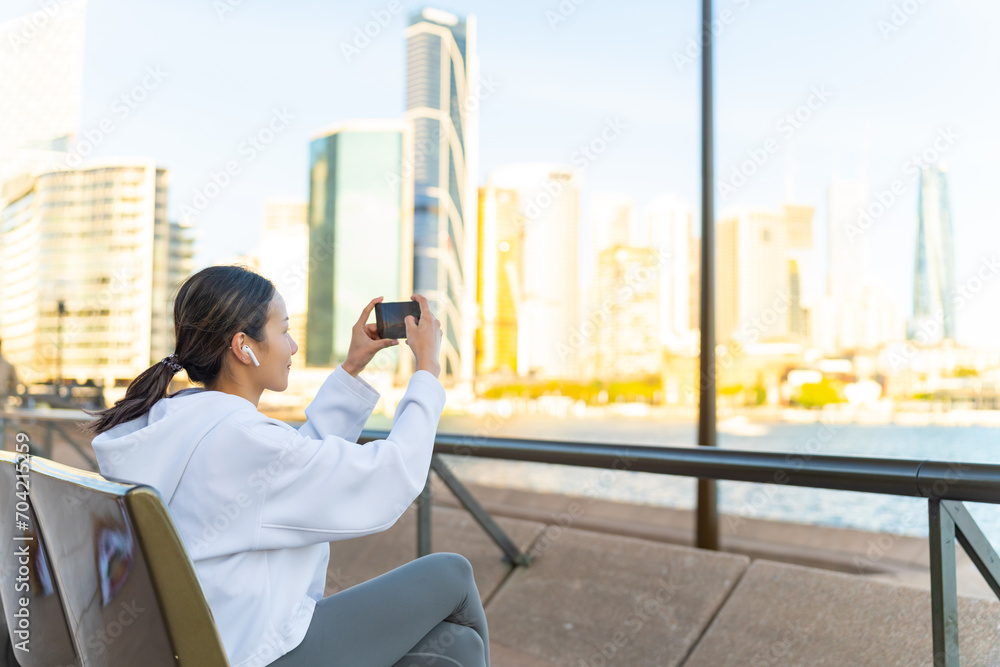 Fototapeta premium Asian woman using smartphone taking picture during jogging exercise in the morning. Attractive girl do sport training running workout in the city. Healthy outdoor lifestyle and technology concept.