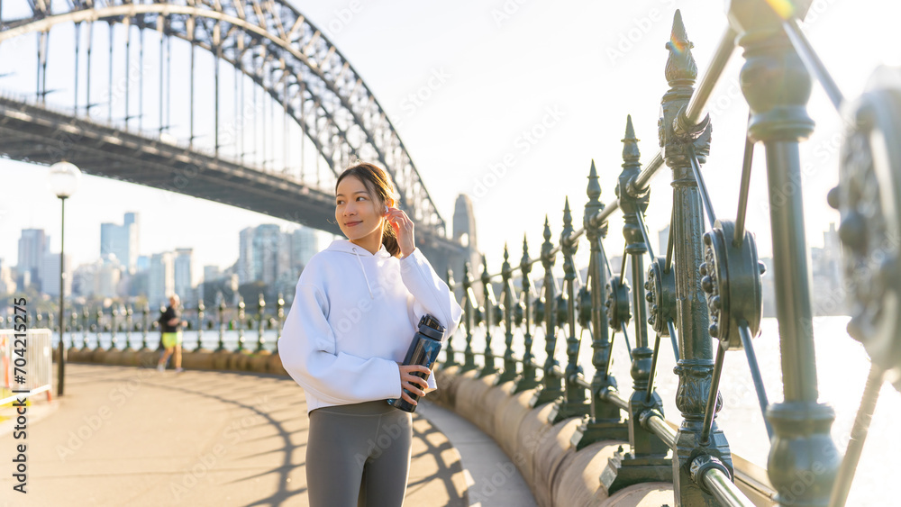 Fototapeta premium Happy Asian woman listening to music from mobile phone and earphones with mobile app during jogging exercise at city street in the morning. Healthy girl do sport training running workout in the city.