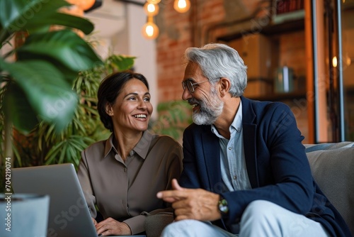 Smiling mature older Indian business man manager having conversation with client or colleague at corporate meeting. Two happy professionals working using laptop and talking sitting, Generative AI 