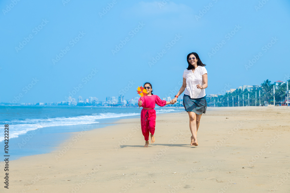Happy Indian mother and daughter running on tropical beach and enjoying summer vacation. daughter playing with paper windmill on sea side.