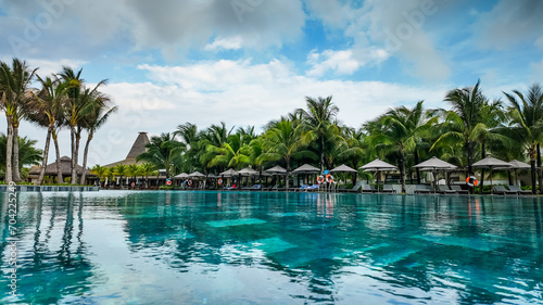 Tropical resort swimming pool with palm trees  sun loungers  and thatched huts under a cloudy sky  conveying a concept of leisure and vacation