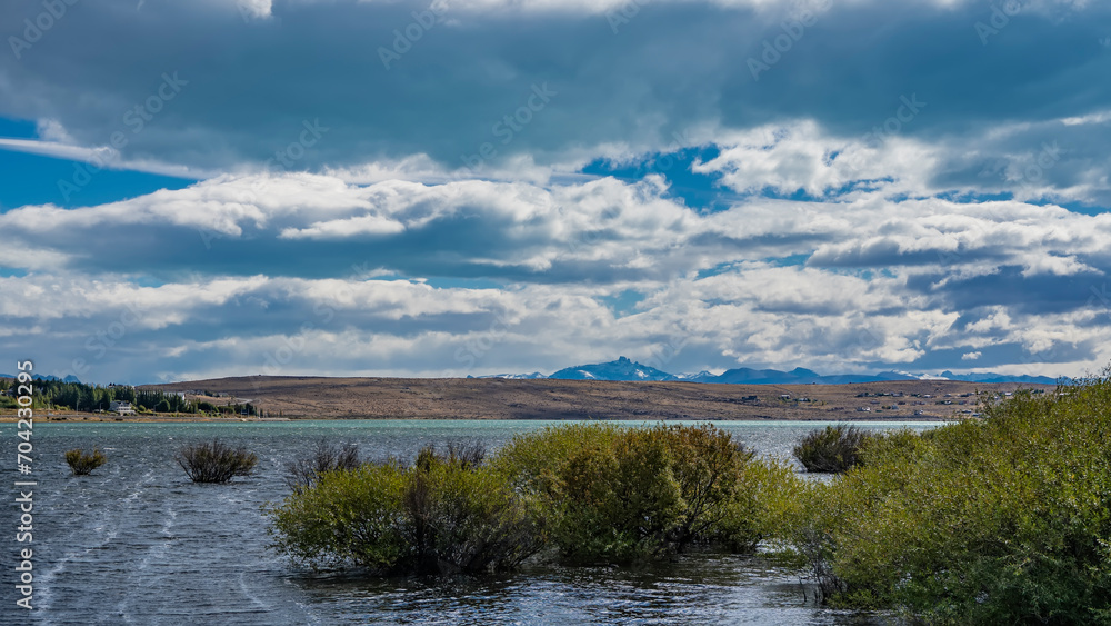 Green bushes grow near the shore of the lake. Ripples on the blue and turquoise water. In the distance, on the coastal hills, houses and roads are visible. Clouds in the sky. Redonda Bay. Argentina. 