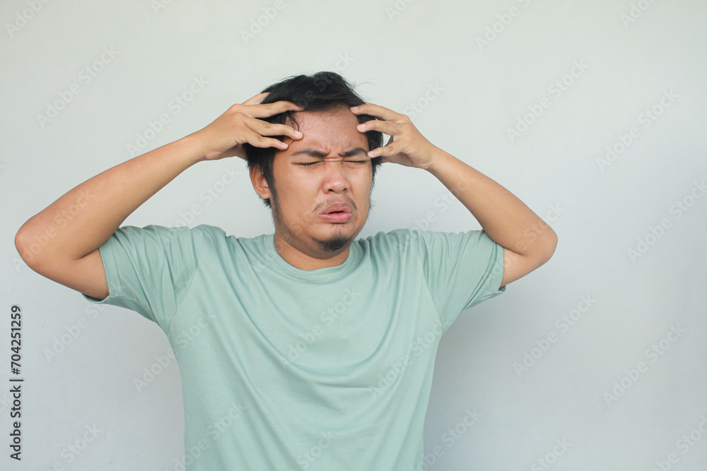 Portrait of young asian man in green t-shirt holding his head with stressed and confused expression. Isolated in gray background. Portrait of young asian man in green t-shirt holding his head with stressed and confused expression. Isolated in gray background.