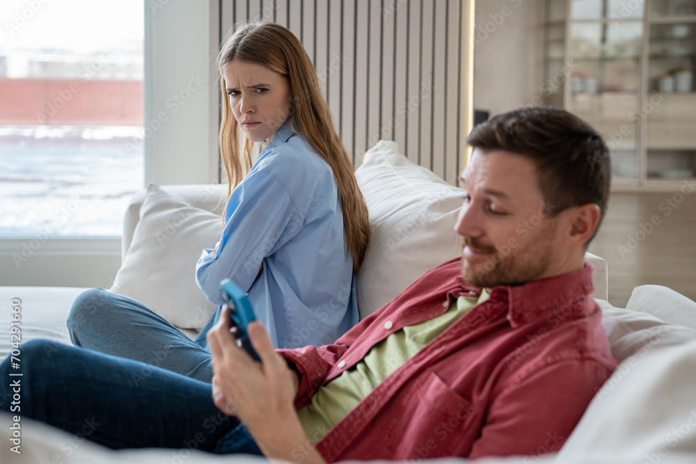 Offended irritated woman looks displeased at husband entertaining with cellphone. Wife gets angry at man obsessed with social media. Female look incredulously at satisfied smiling boyfriend with phone