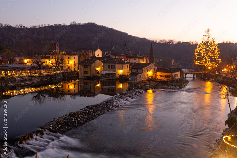 Festive Lights Reflect: Christmas 2024 in Borghetto sul Mincio. Bright reflections on calm waters create a enchanting nocturnal spectacle of celebration and magic