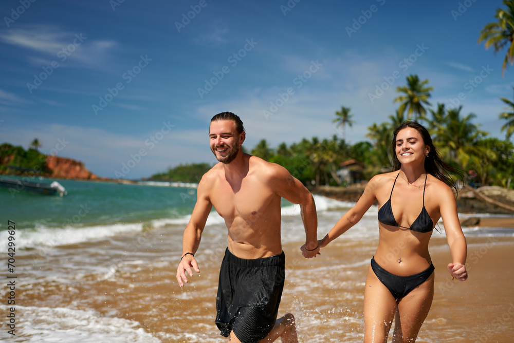 Joyful couple runs on tropical beach, hand-in-hand. Athletic man and slender woman exercise by sea. Fit pair enjoys sunny vacation, laughs during scenic seaside sprint. Outdoor activity, health, fun. Joyful couple runs on tropical beach, hand-in-hand. Athletic man and slender woman exercise by sea. Fit pair enjoys sunny vacation, laughs during scenic seaside sprint. Outdoor activity, health, fun.