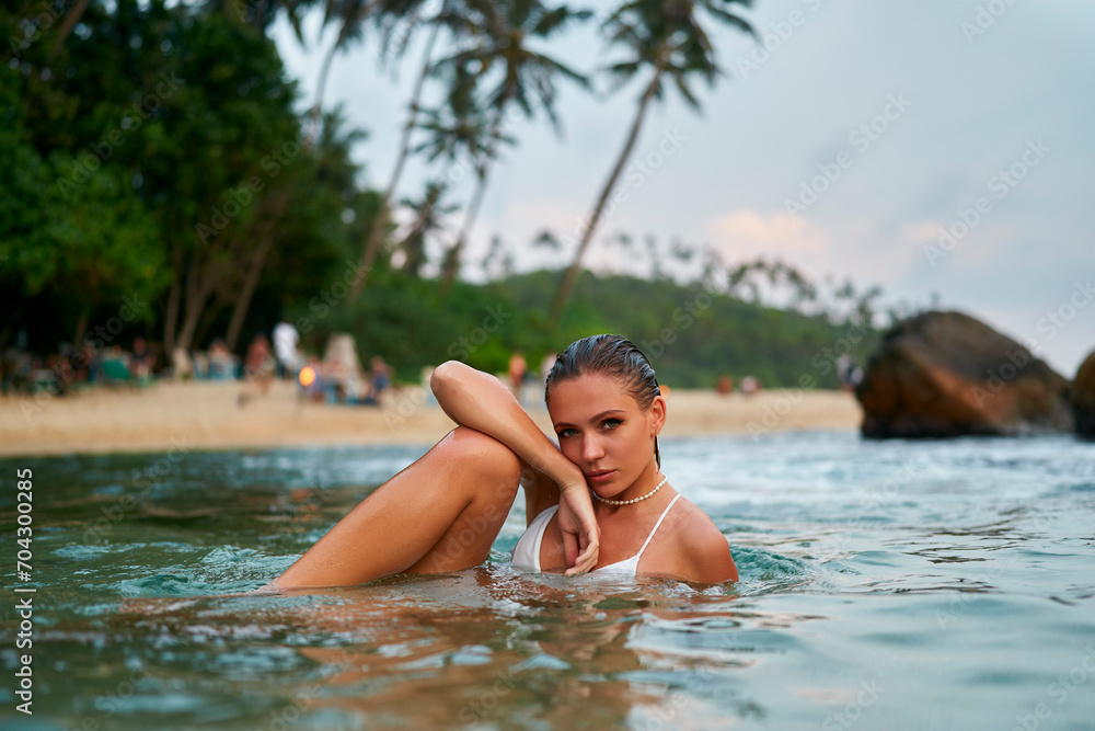 Woman lounges in ocean water, enduring makeup intact, tropical beach backdrop. Traveler enjoys serene sea, durable cosmetics, fashion display in nature. Holiday, relaxation, style, durability merge.
