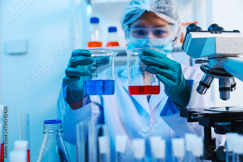 Asian people scientist in lab coat and protective gloves working with test tubes with green and red liquids, with microscope and other test tubes in the background in laboratory.