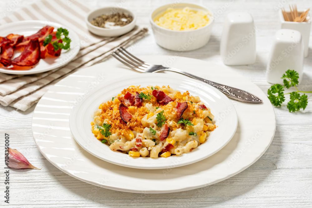 portion of mac and cheese with corn, bacon topped with panko breadcrumbs on white plate on white wooden table with ingredients at background, horizontal view from above, close-up portion of mac and cheese with corn, bacon topped with panko breadcrumbs on white plate on white wooden table with ingredients at background, horizontal view from above, close-up