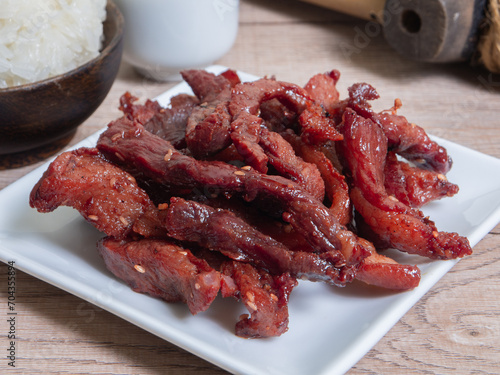  fried sun dried pork in a ceramic dish served with sticky rice on wooden table. thai style delicious food menu.