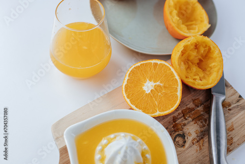There is a manual juicer on a wooden cutting board  next to it is a knife and half a squeezed orange. a glass of juice and a plate of oranges. White background
