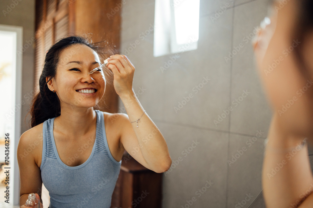 Cute smiling Asian young woman applies moisturizing serum from pipette to her face while standing in front of mirror. Lovely charming Korean girl taking care of her skin. Cute smiling Asian young woman applies moisturizing serum from pipette to her face while standing in front of mirror. Lovely charming Korean girl taking care of her skin.