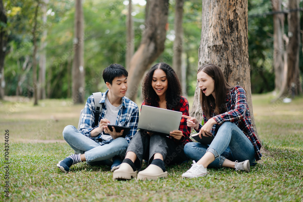 Three young college students is reading a book while relaxing sitting on grass in a campus park with her friends. Education concept