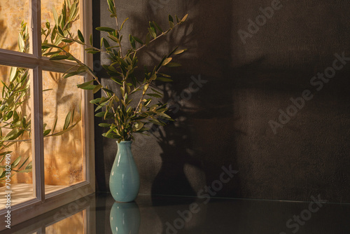 Panorama de fond d'un mur noir avec un vase et des branches d'oliviers pour création d'arrière plan. Panoramique avec jeu d'ombre et de lumière à travers une fenêtre.	 photo