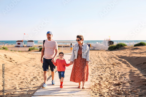 Happy family, mother father and little daughter, walking towards summer sea beach along wooden path