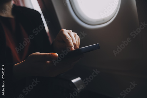 Blonde female tourist checking incoming notification on smartphone sitting on seat of airplane with netbook.Young businesswoman share media from telephone on laptop computer during plane flight photo
