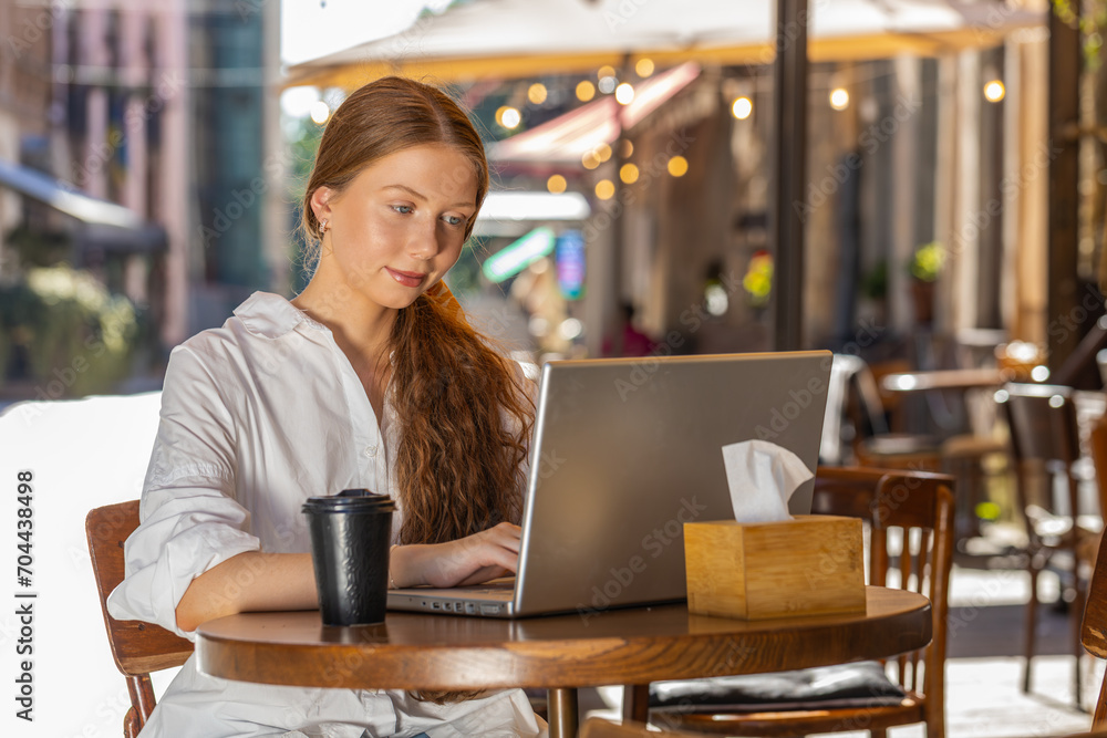 Fototapeta premium Redhead child girl working study education online distant job with laptop in city sunny street cafe restaurant browsing website chatting outdoors during break. Smiling woman tourist freelancer