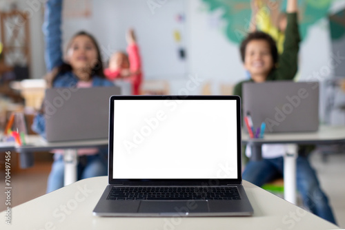 Laptop computer with blank mockup screen on teachers table with junior children pupils raising hands in classroom. Education software website technology ads concept