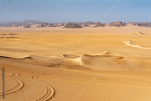 Landscape of Erg Admer in the Sahara desert, Algeria. The golden sand of Erg Admer with, in the distance, the rock formations of Tassilis. In the foreground, we can see the tracks left by jeep tires