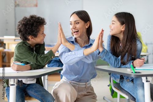Two pupils and female teacher cheering, giving high five to each other and smiling, enjoying time after lesson at school, satisfying of result