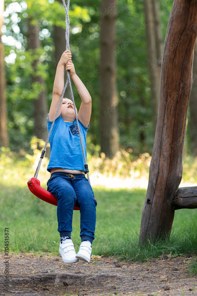 Portrait of a dreamy boy sitting on a swing with a red seat. The child twists the ropes and quickly spins around its axis backwards. An interesting way to swinging