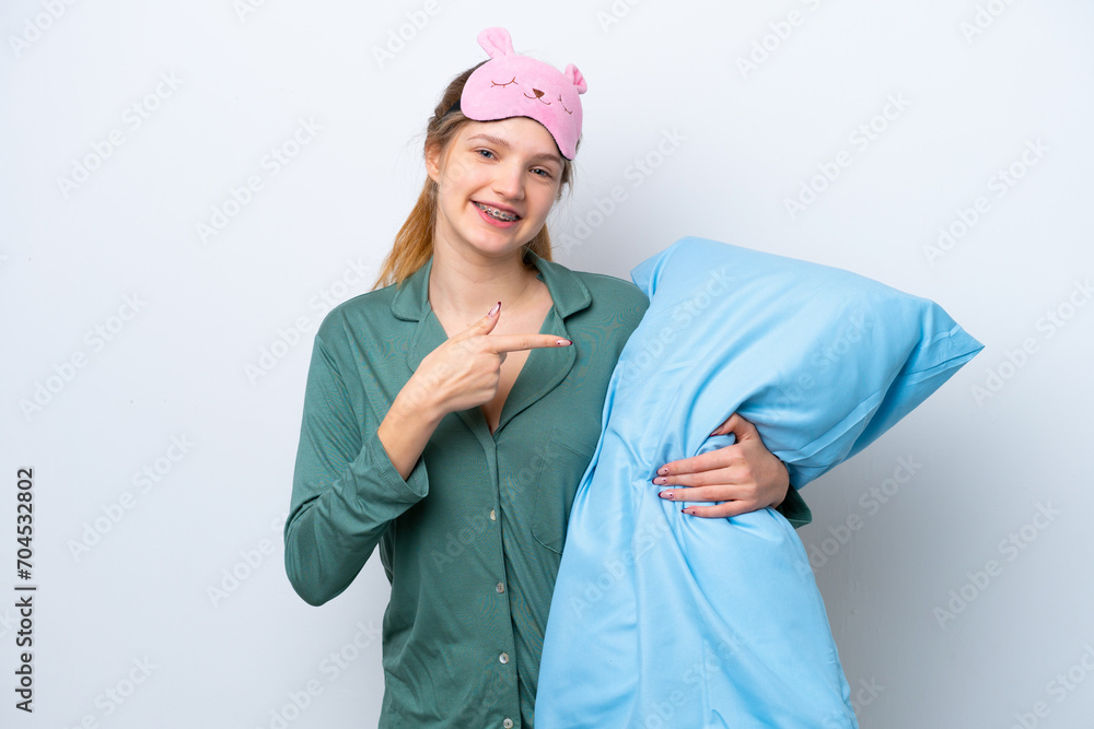 Teenager Russian girl isolated on white background in pajamas and pointing a pillow Teenager Russian girl isolated on white background in pajamas and pointing a pillow