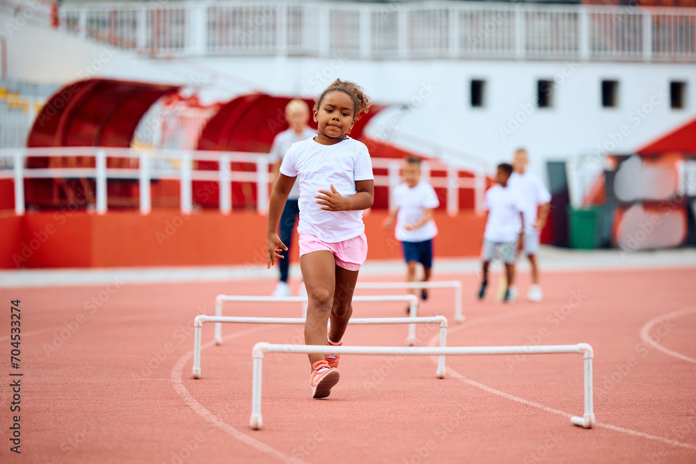 Black little girl on running track with hurdles at stadium.