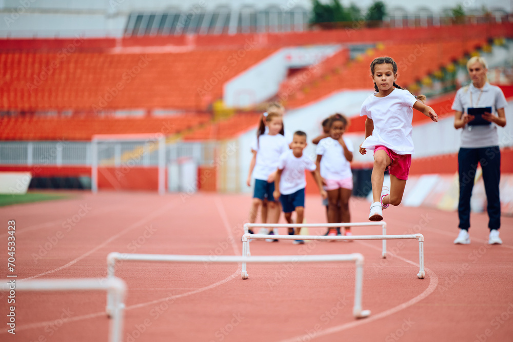 Little girl jumping over hurdle during PE class at stadium.