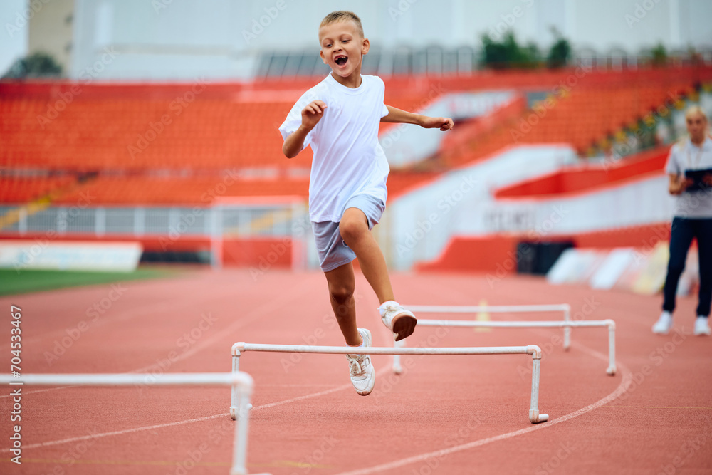 Cheerful kid umping over obstacles while running during sports training at stadium.
