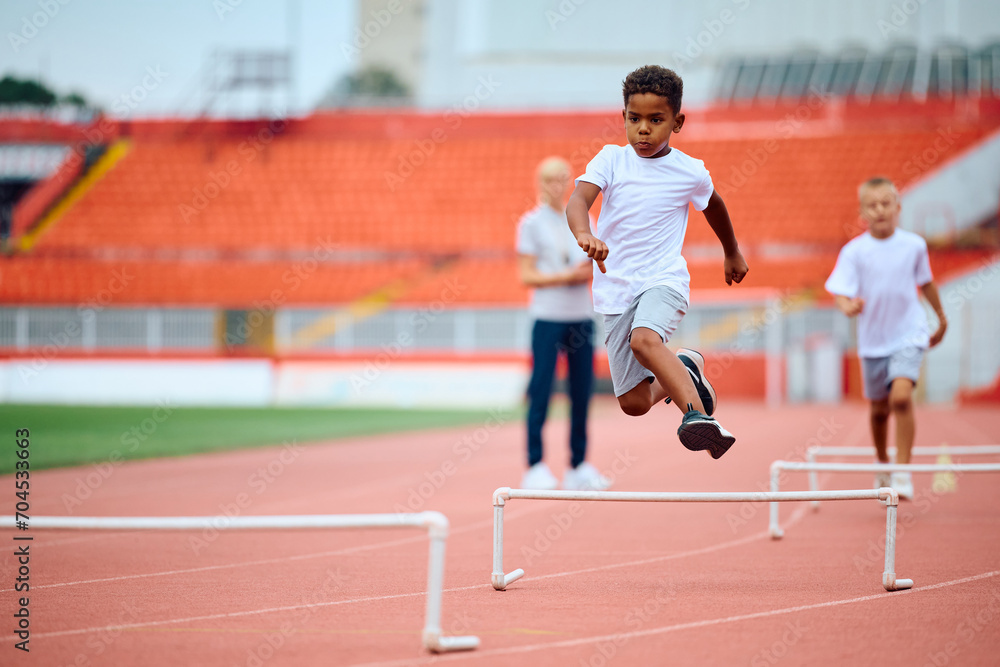 Black kid jumping over hurdles while running during exercise class at stadium.
