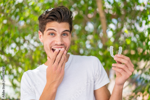 Young caucasian man at outdoors holding invisible braces with surprised expression