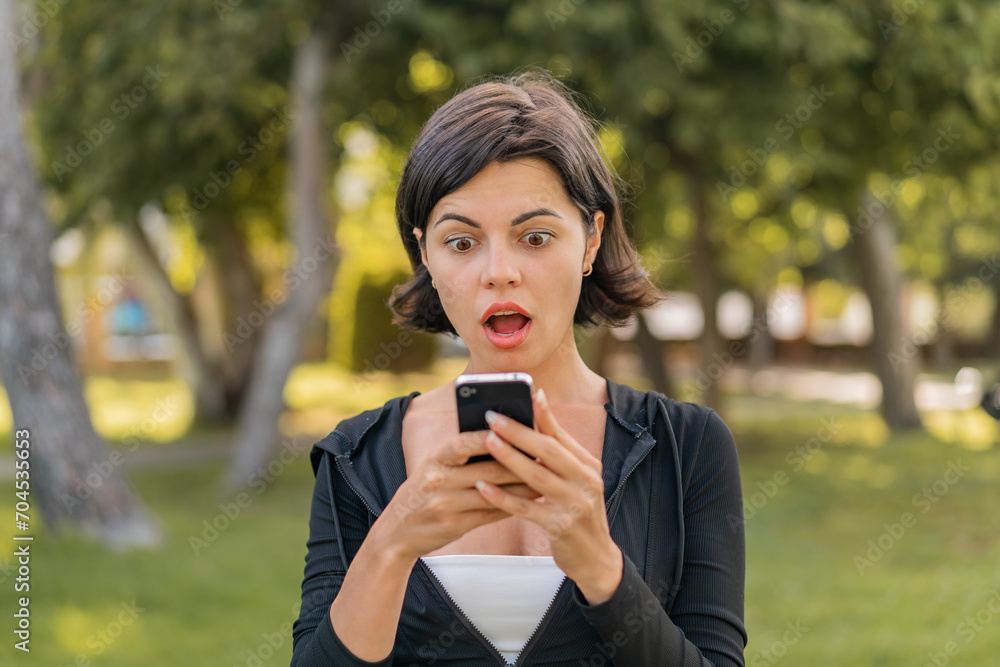 Young pretty Bulgarian woman at outdoors looking at the camera while using the mobile with surprised expression