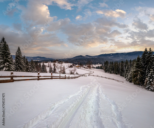 Secondary countryside alpine road in remote mountain village, snow drifts and wood fence on wayside