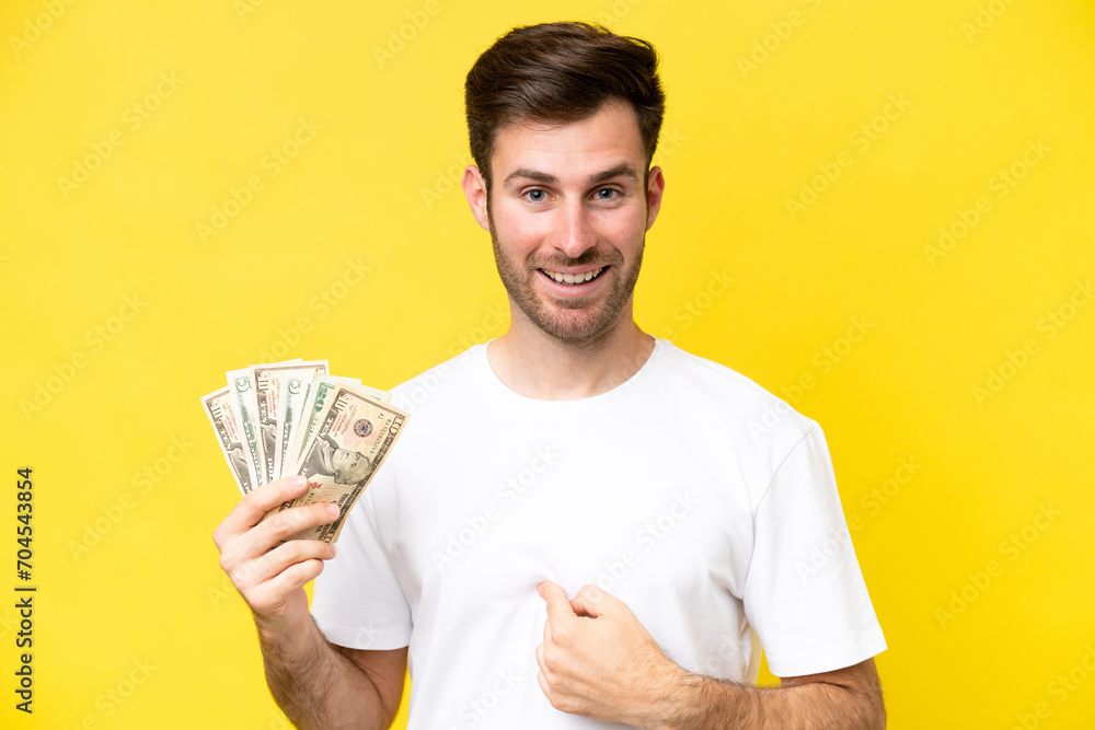 Young caucasian man taking a lot of money isolated on yellow background with surprise facial expression Young caucasian man taking a lot of money isolated on yellow background with surprise facial expression