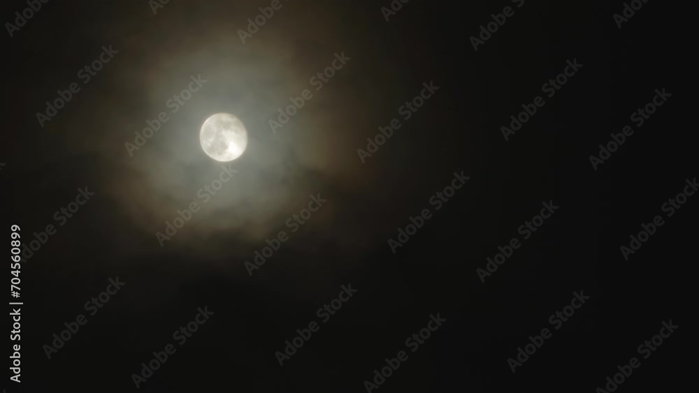 Full moon on a dark cloudy night with colored clouds flying in the sky in front of the Moon. Corona - light optical phenomenon Full moon on a dark cloudy night with colored clouds flying in the sky in front of the Moon. Corona - light optical phenomenon