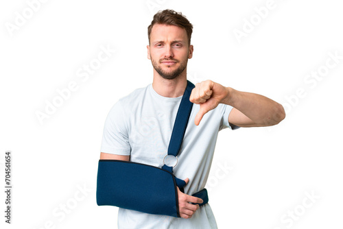 Young caucasian man with broken arm and wearing a sling over isolated chroma key background showing thumb down with negative expression
