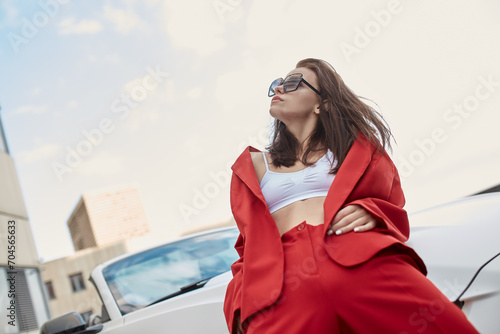 Relaxed young woman in sunglasses and red pantsuit leaning against hood of convertible