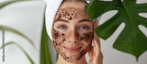 A youthful, pretty girl with a towel wrapped on her head applies a coffee scrub on her face and holds a leaf.