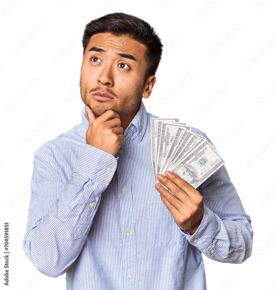 Young Chinese man holding dollar bills in studio looking sideways with doubtful and skeptical expression. Young Chinese man holding dollar bills in studio looking sideways with doubtful and skeptical expression.