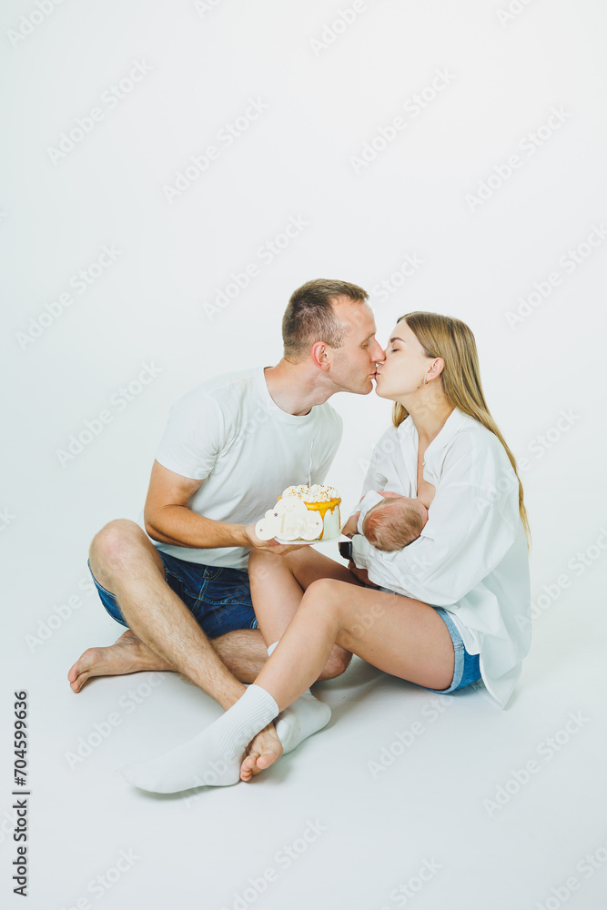 Young happy parents with a baby in their arms and a cake celebrate the first month of the baby's life. Mom and dad with a newborn baby on a white background Young happy parents with a baby in their arms and a cake celebrate the first month of the baby's life. Mom and dad with a newborn baby on a white background