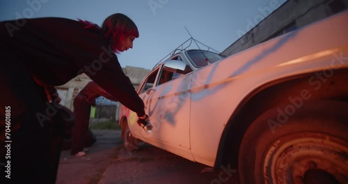 Post-apocalypse. Riot Fire. Protest. Vandalism Punk protester girl uses a red pressure paint finishing vandal graffiti riot ugly lettering on viciously crushed old car on the street